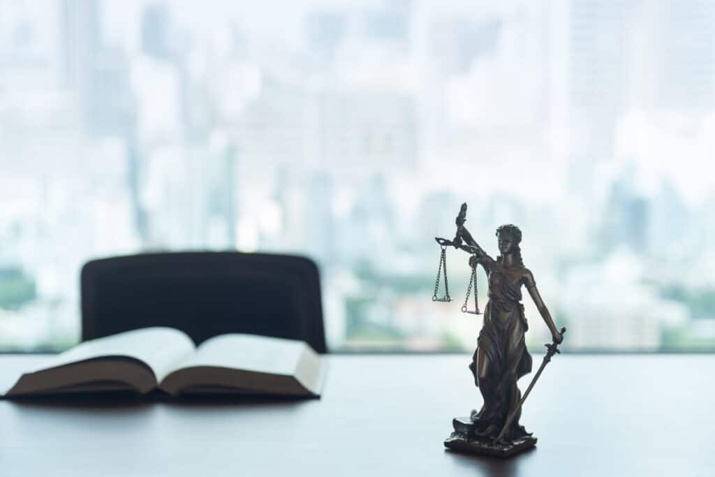 Bronze Lady Justice statue holding scales and a sword on a desk beside an open book, with a blurred cityscape in the background conveys law and justice.